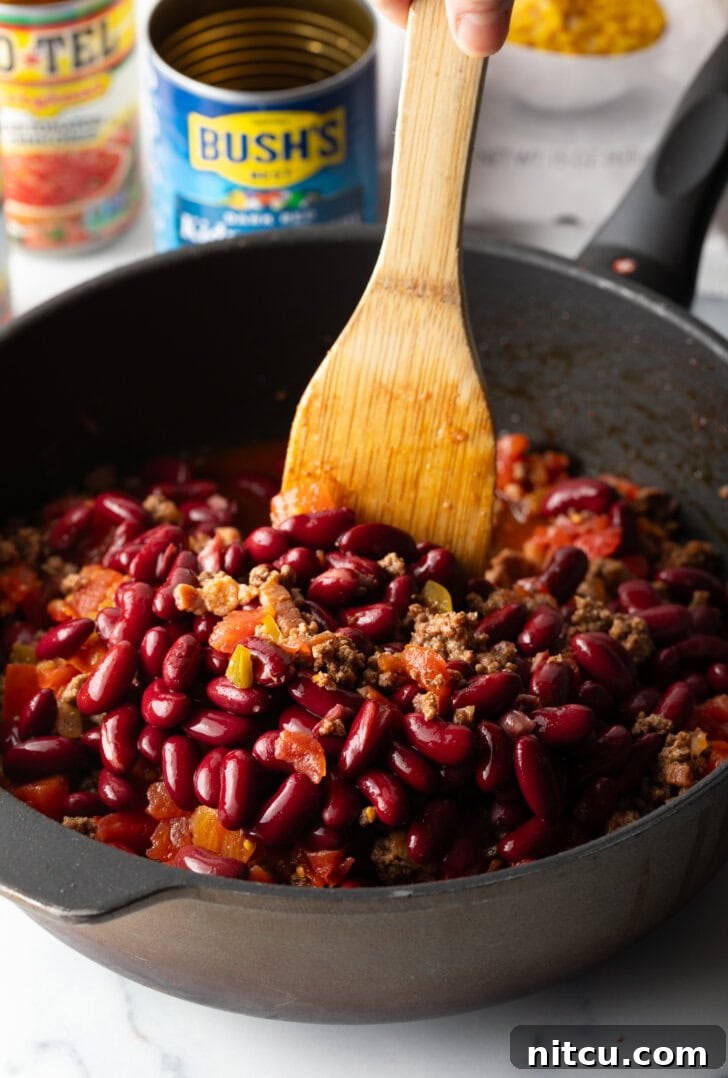 Deep black skillet with ground beef mixture and beans, being stirred by a wooden spatula.