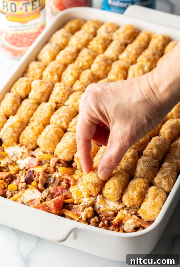 Hand adding frozen tater tots to the top of a baking dish casserole.