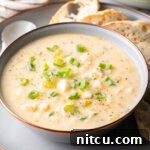 Chowder in a white bowl, on a gray plate surrounded by slices of bread and a spoon t the side.
