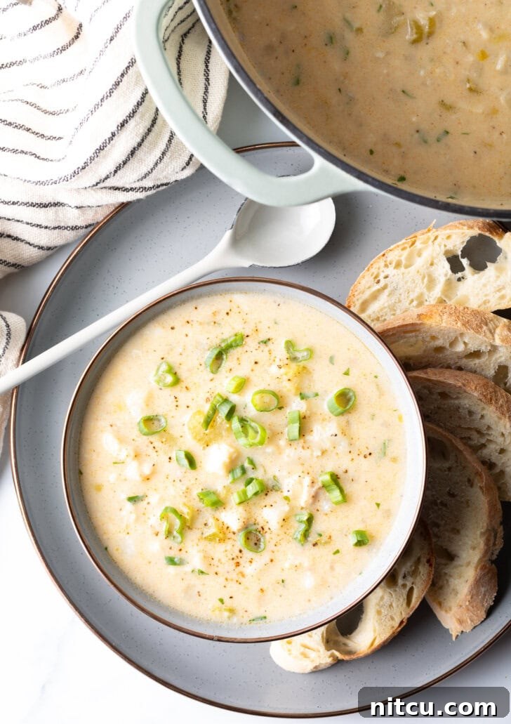 Top down view of the chowder in a white bowl, on a gray plate surrounded by slices of bread and a spoon t the side.