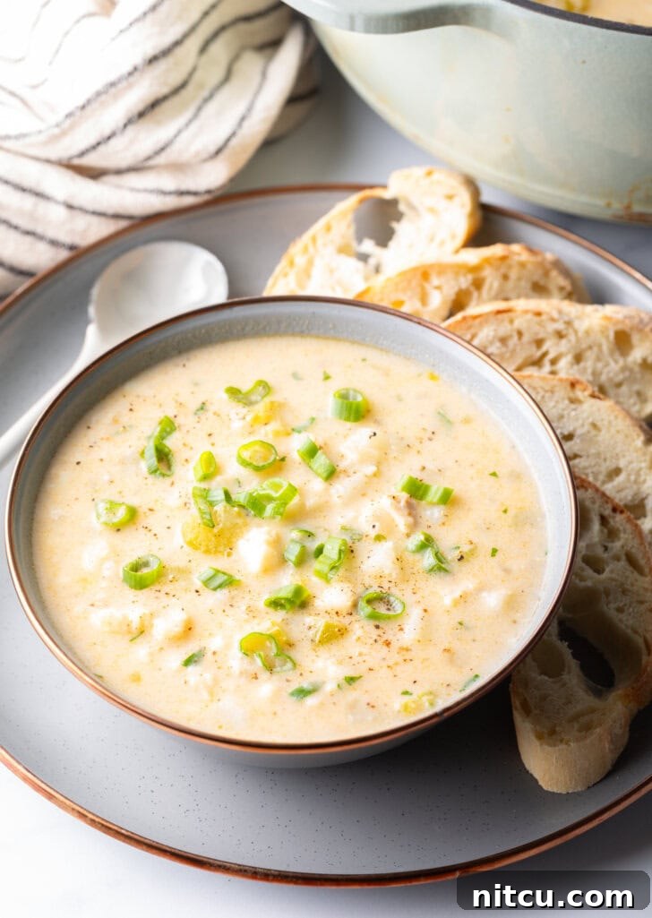 Fish Chowder in a white bowl, on a gray plate surrounded by slices of bread and a spoon t the side.