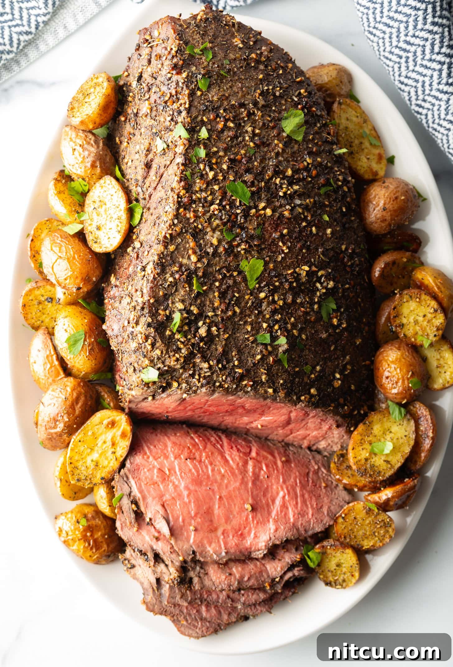 Cooked beef round roast on a metal baking pan. The front half of the roast has been thinly sliced and the meat is surrounded by roasted baby potatoes.
