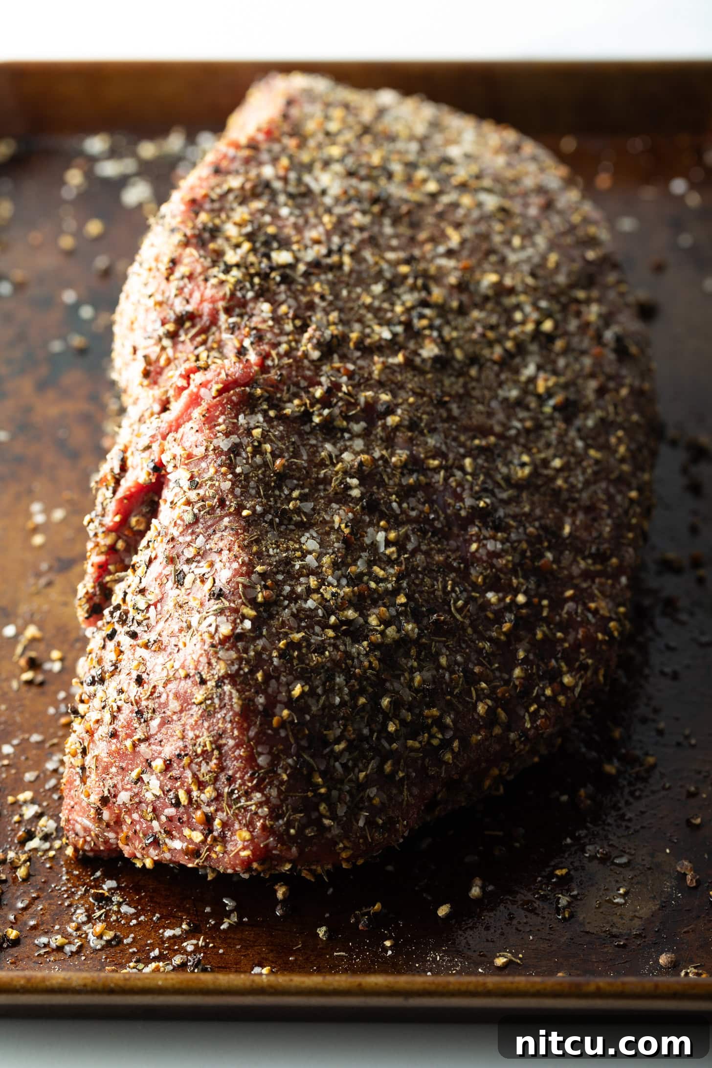 Raw top round roast coated with pepper rub, on a baking sheet.