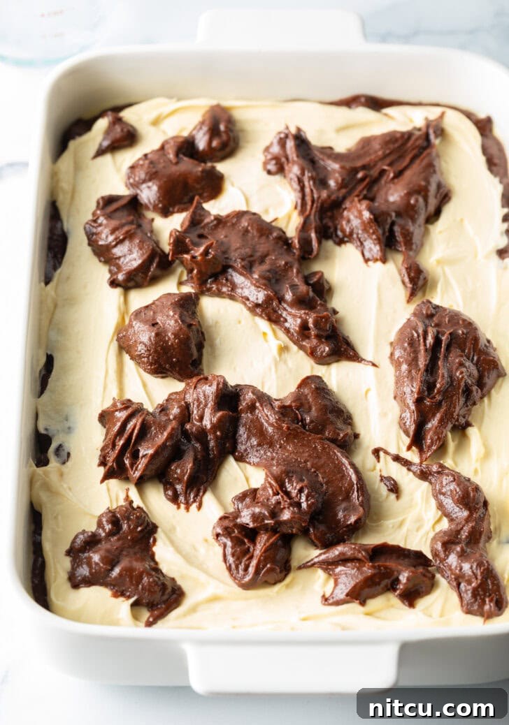 A top-down view of a white rectangular baking pan filled with layers of brownie batter, topped with cream cheese, and further dotted with dollops of more brownie batter, ready for swirling.