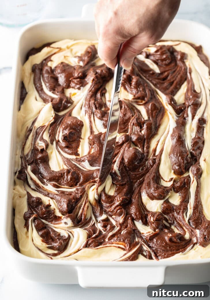 A hand uses a knife to artfully swirl the raw cheesecake and brownie batter together, creating a beautiful marbled pattern.
