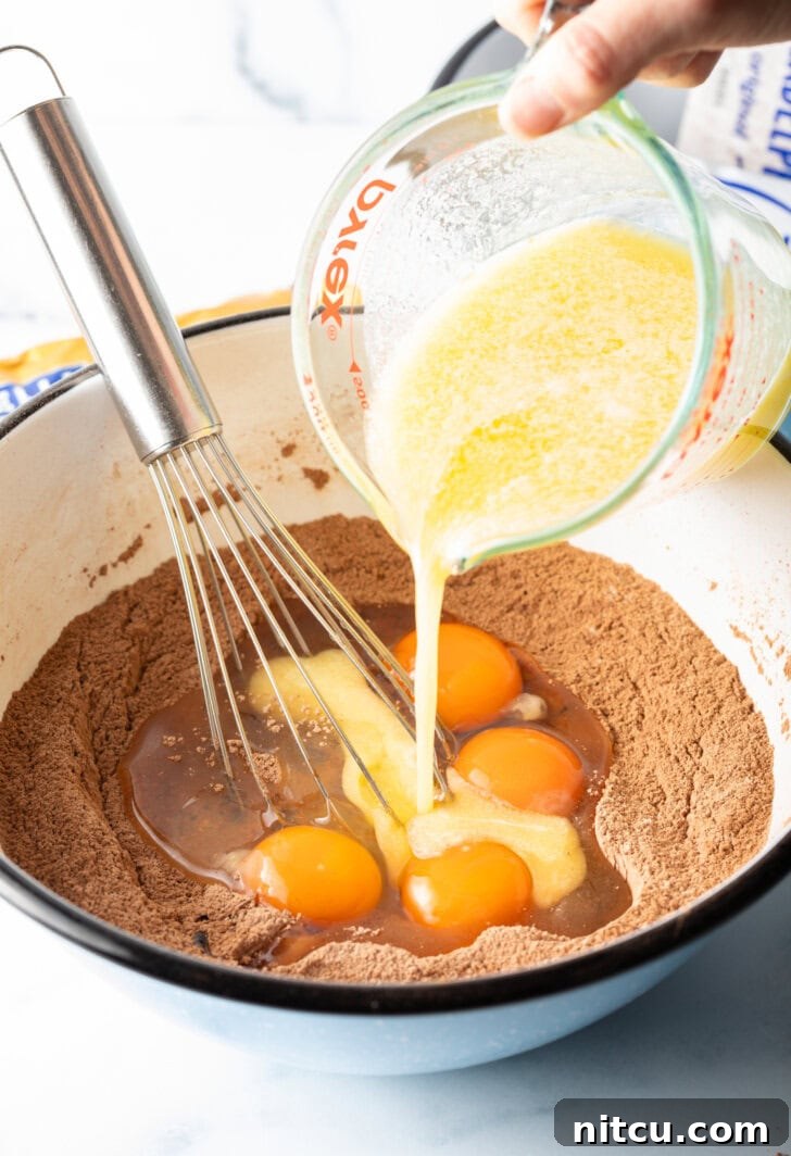 Melted butter is being poured from a glass measuring cup into a white bowl containing dry ingredients and cracked eggs, ready to be mixed into brownie batter.