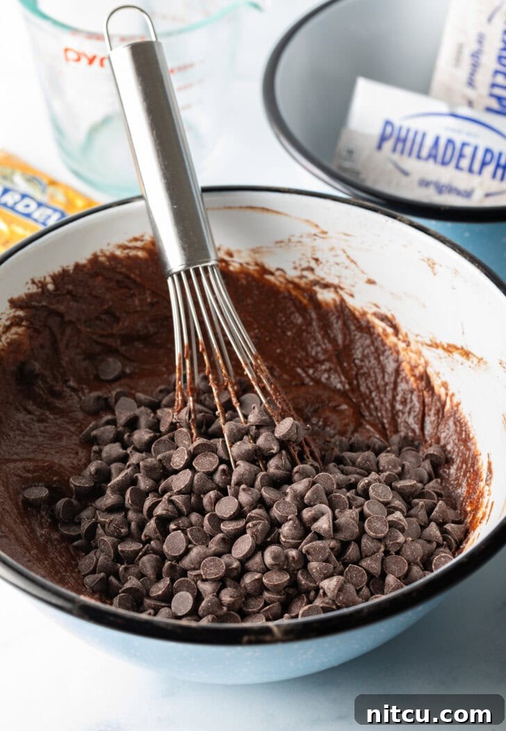 A whisk is gently incorporating chocolate chips into rich brownie batter in a pristine white mixing bowl.