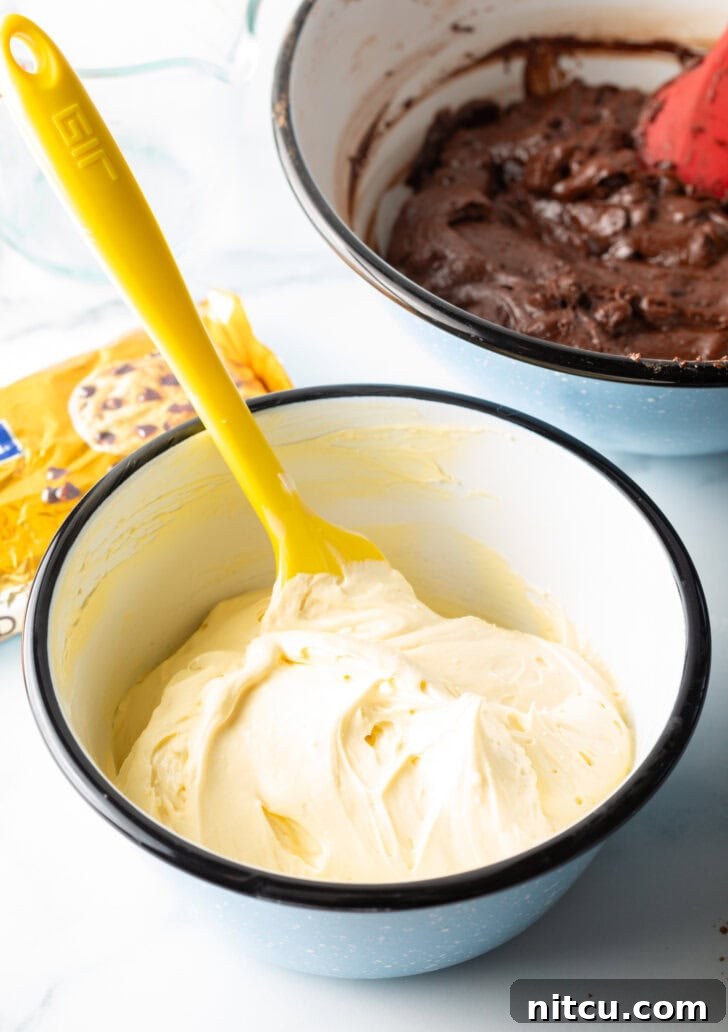 A yellow spatula is stirring a smooth, creamy cream cheese filling in a small white bowl, ready for layering.