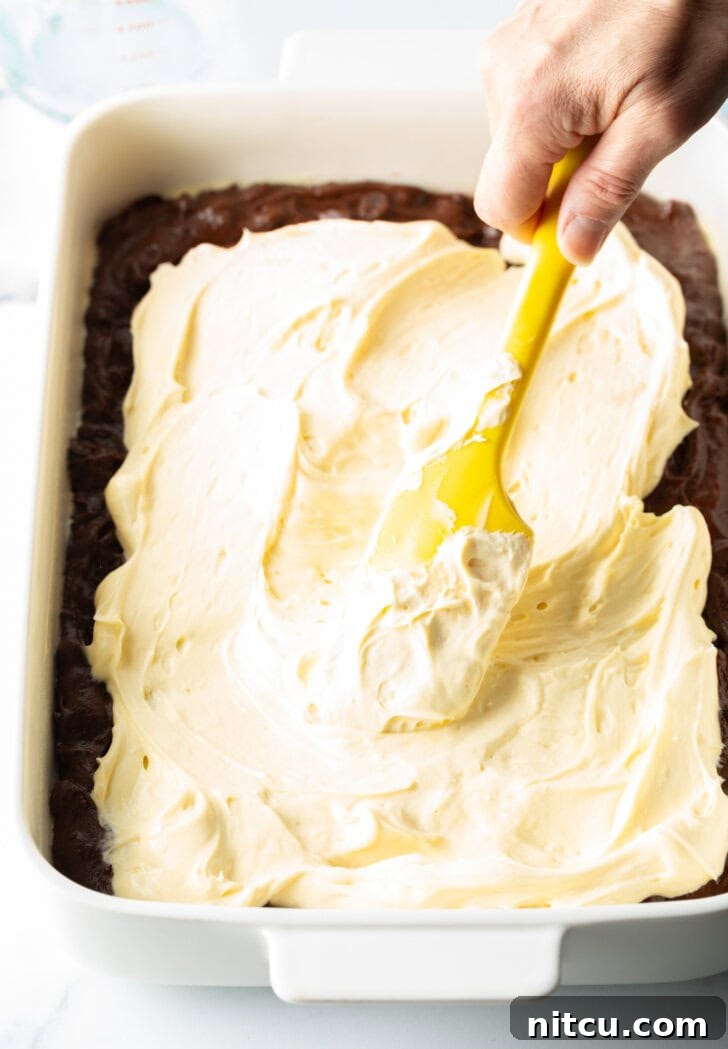 A hand uses a yellow spatula to gently spread a creamy cheesecake filling over the top of a layer of brownie batter in a baking pan.