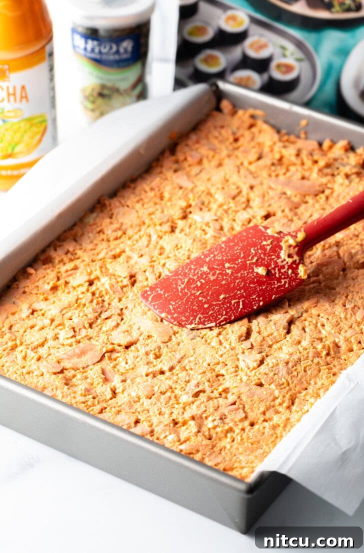 A red spatula spreading the mixed salmon and sriracha mayonnaise mixture evenly over the cream cheese layer in the baking pan.