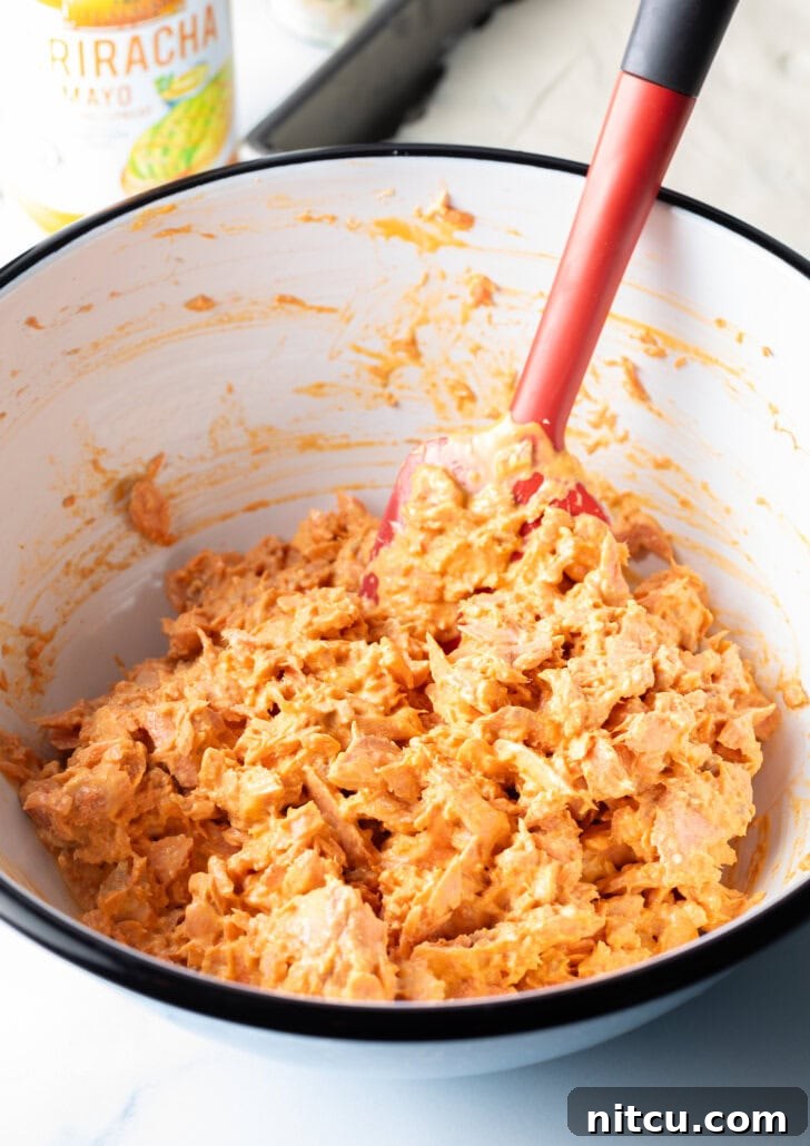 Close-up of cooked salmon being flaked and mixed with sriracha mayonnaise in a white bowl using a red spatula, creating the flavorful topping.