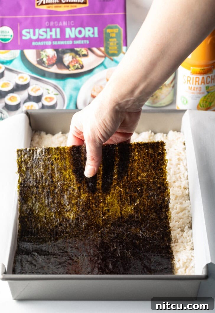 A hand carefully placing a large sheet of nori seaweed on top of the pressed rice layer in a rectangular baking pan.