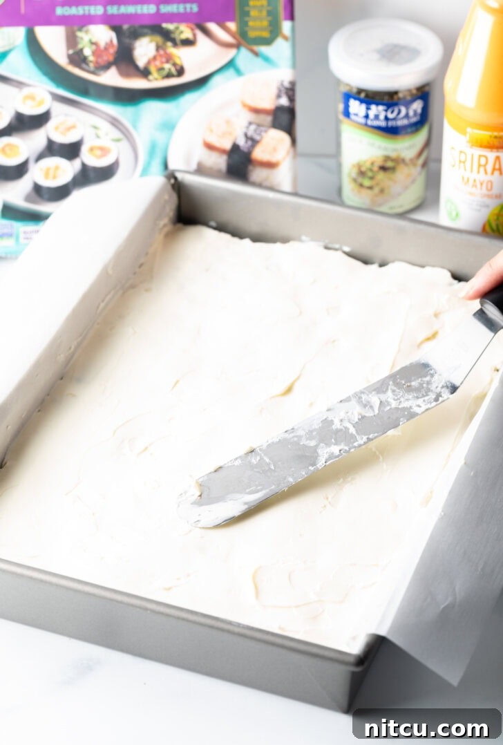 A flat metal spatula smoothly spreading softened cream cheese over the nori layer in the baking pan, creating a creamy white layer.