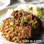 Two slices of meatloaf on a plate with a scoop of mashed potatoes, all topped with chopped green onions for garnish.