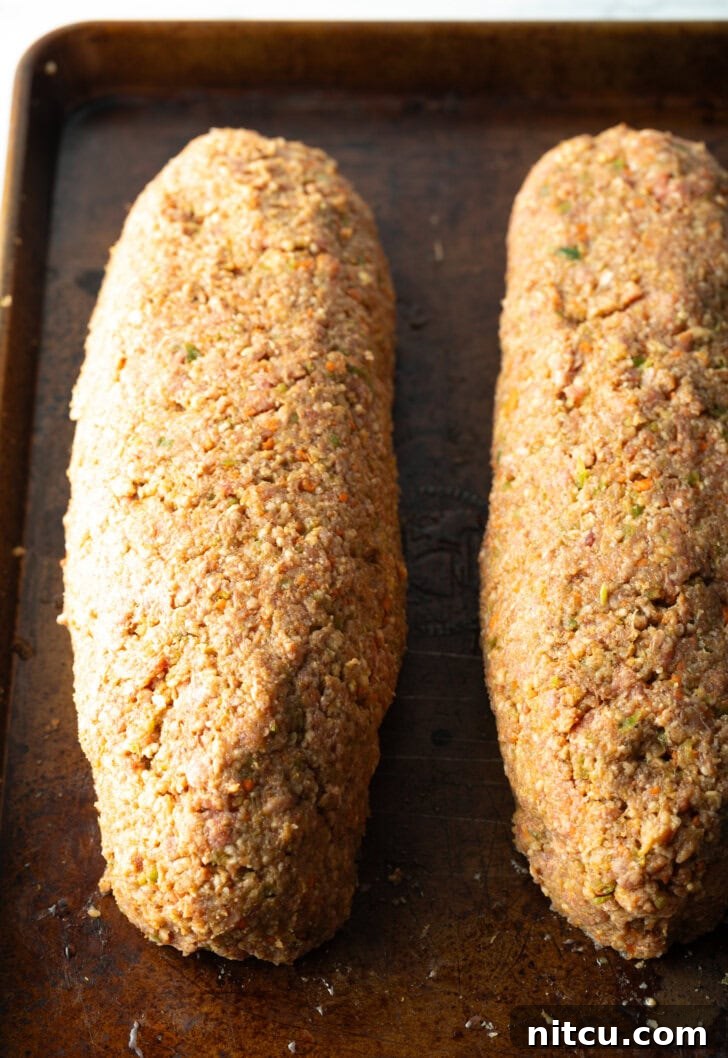 Two raw, perfectly shaped meatloaves resting side-by-side on a baking sheet, glistening with a coat of barbecue sauce, ready for the smoker.