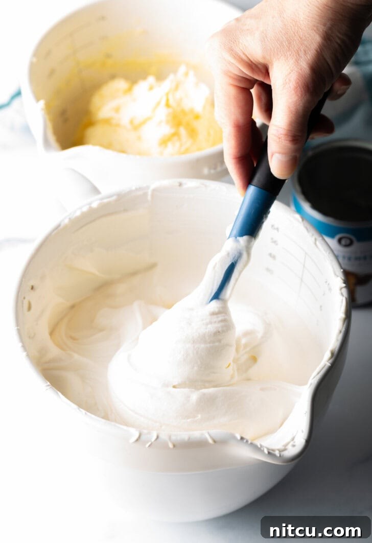 A hand gently blending a creamy vanilla ice cream base in a large white mixing bowl using a blue rubber spatula, demonstrating the delicate folding technique.