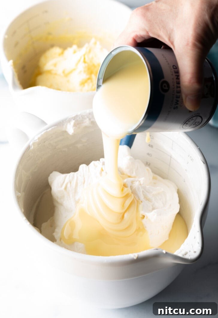 A hand pouring a stream of luscious sweetened condensed milk over the gently whipped ingredients in a mixing bowl, an essential step for no-churn ice cream.