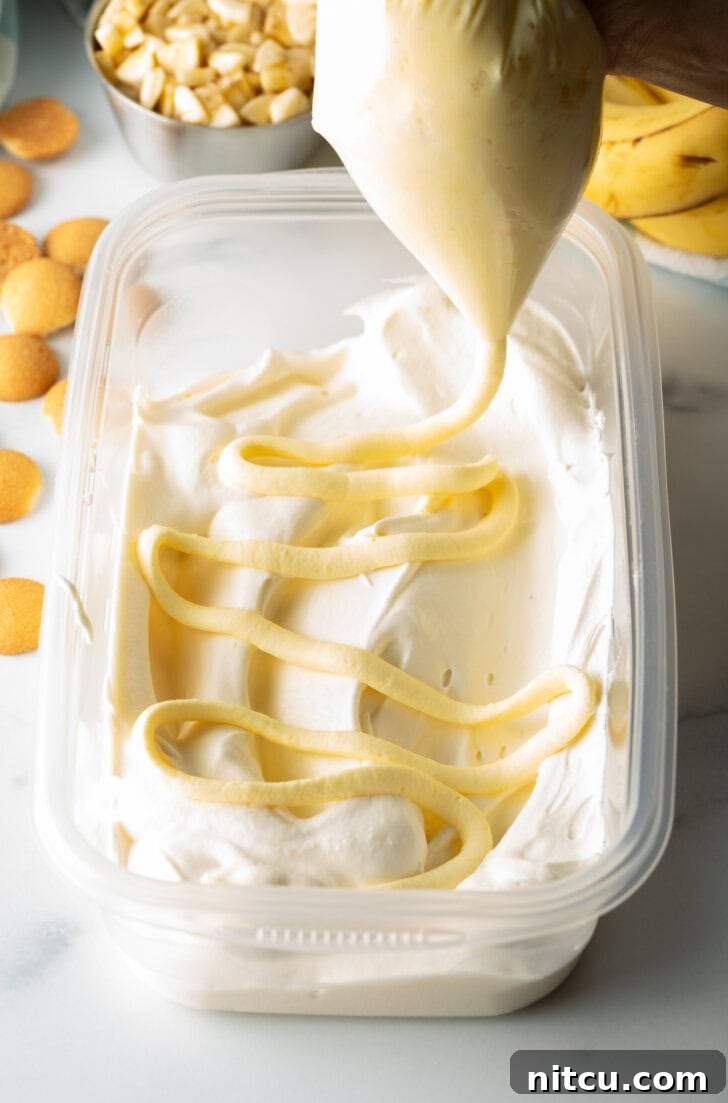 A hand expertly squeezing rich banana pudding from a piping bag onto a layer of frozen ice cream within a rectangular freezer container, creating a beautiful swirl.