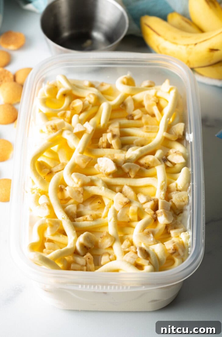 Perfectly arranged slices of ripe banana nestled within a rectangular freezer container, layered among the creamy ice cream base, awaiting the next delicious addition.