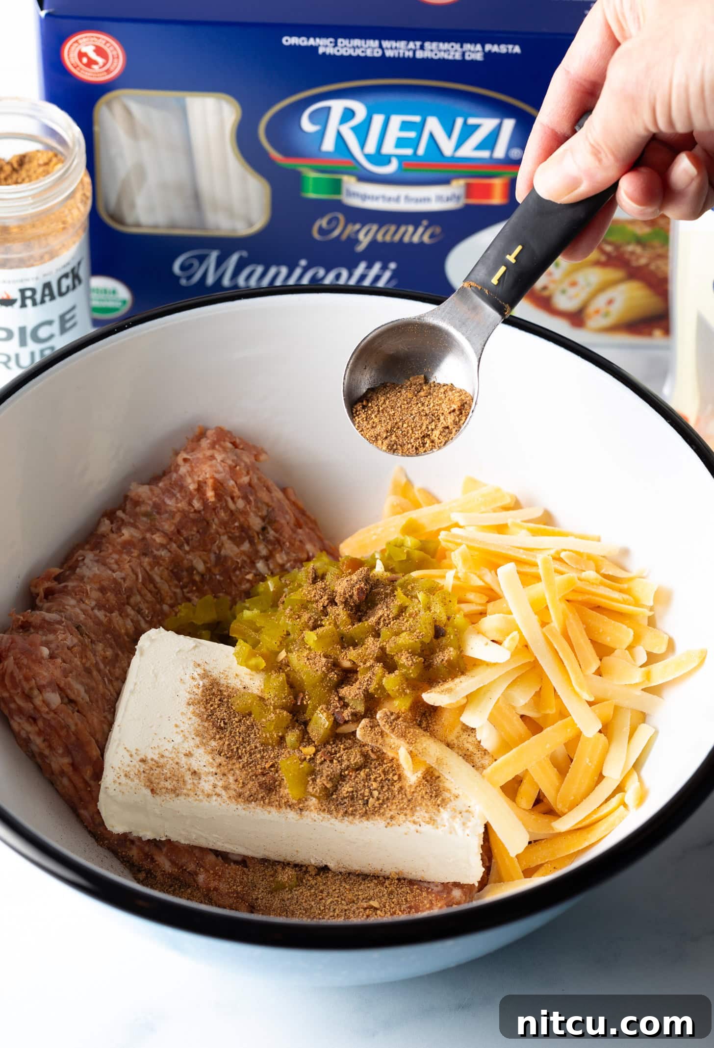 Hand adding tablespoon of seasoning to a white bowl with raw ground beef, shredded cheddar cheese, block of cream cheese, and chopped peppers.