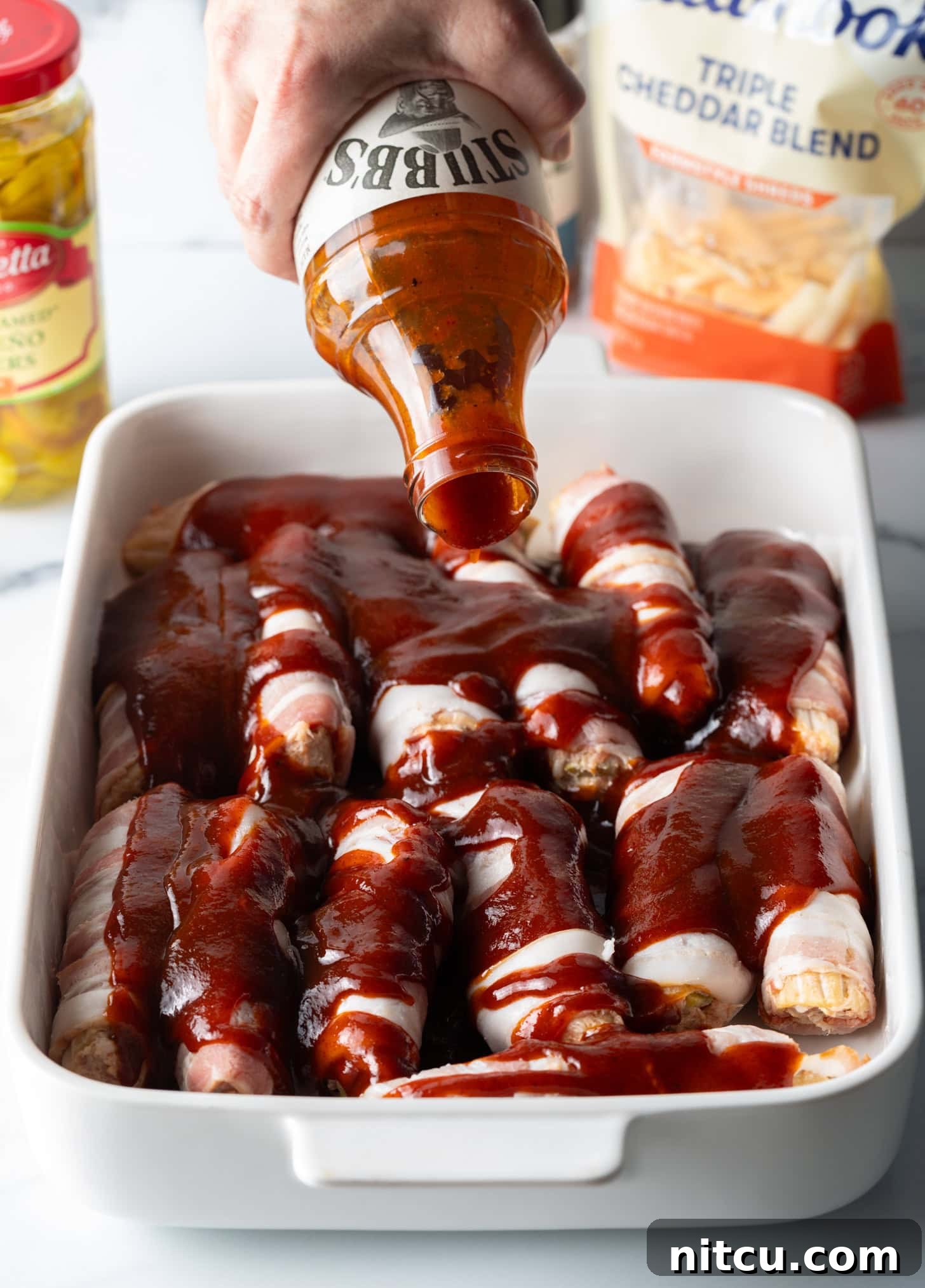 Hand pouring barbecue sauce from a bottle on top of the bacon wrapped and stuffed shells layered in a white baking dish.