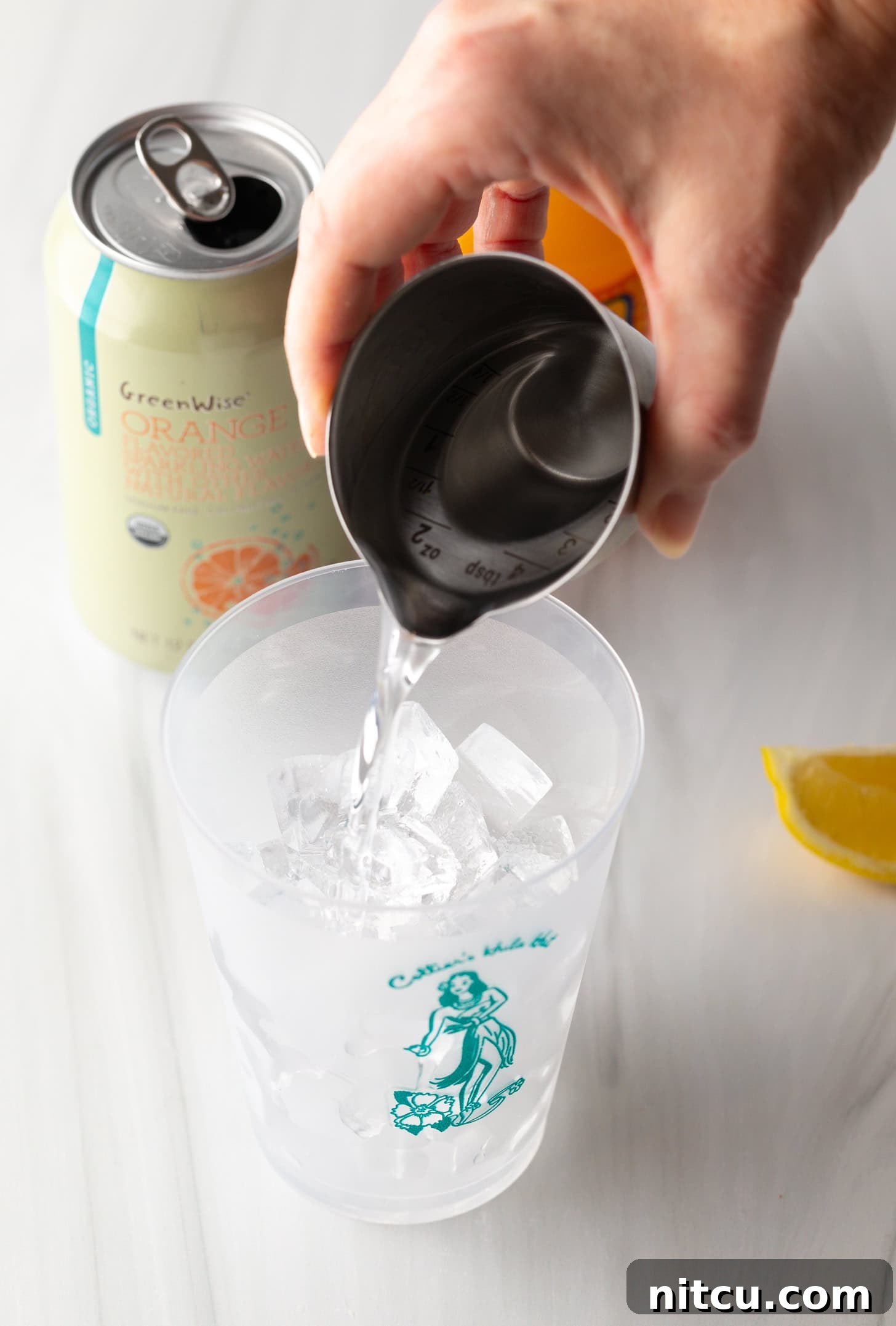 Hand pouring vodka from a metal shot glass into a glass filled with ice.