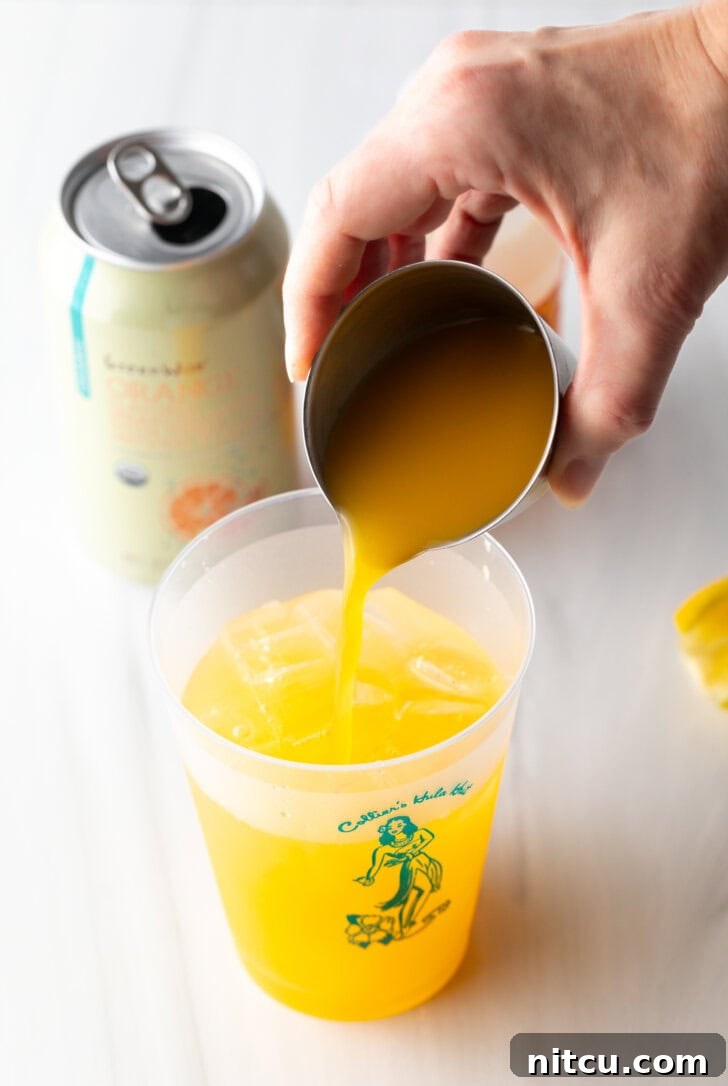Hand adding orange juice from a metal shot glass into a glass cup.
