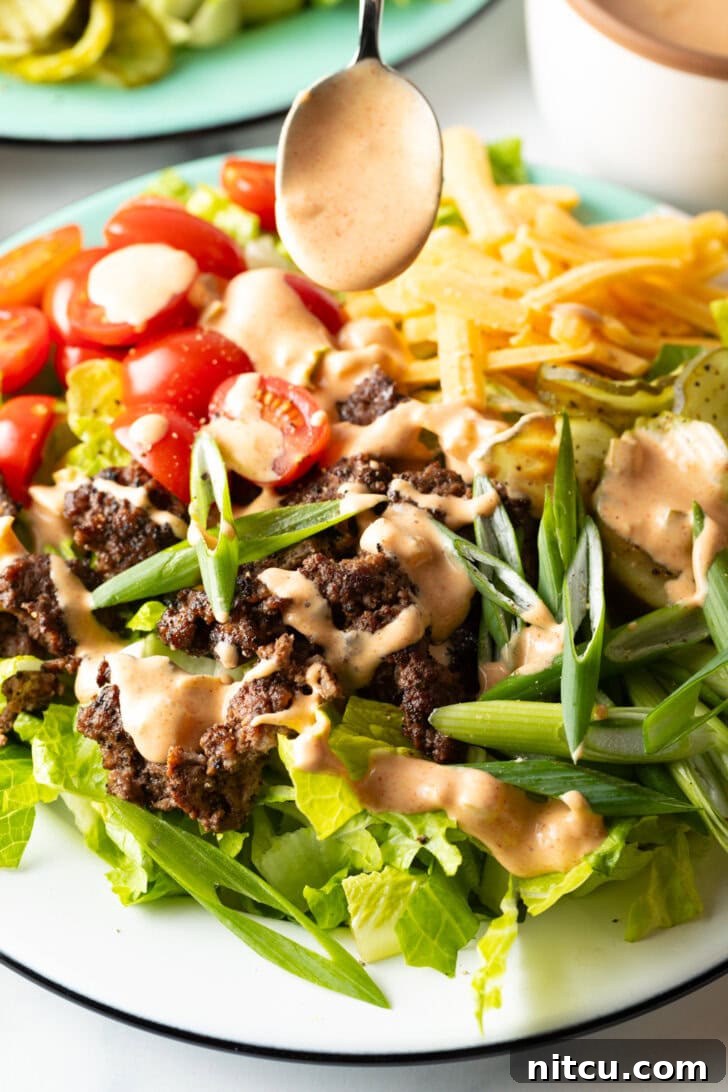 A spoon generously drizzling creamy, golden Big Mac sauce over the loaded burger salad, enhancing its appeal and flavor.