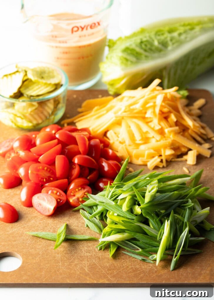 Recipe ingredients on a cutting board: Neat piles of bright shredded cheese, halved red cherry tomatoes, vibrant sliced green onions, and a small glass brimming with crisp pickle slices, highlighting fresh preparation.
