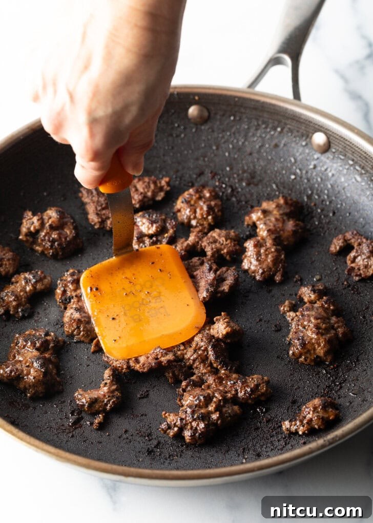 A hand with a spatula expertly cooking mini burger patties in a hot skillet, ensuring a perfect sear and delicious browned crumbles for the salad.