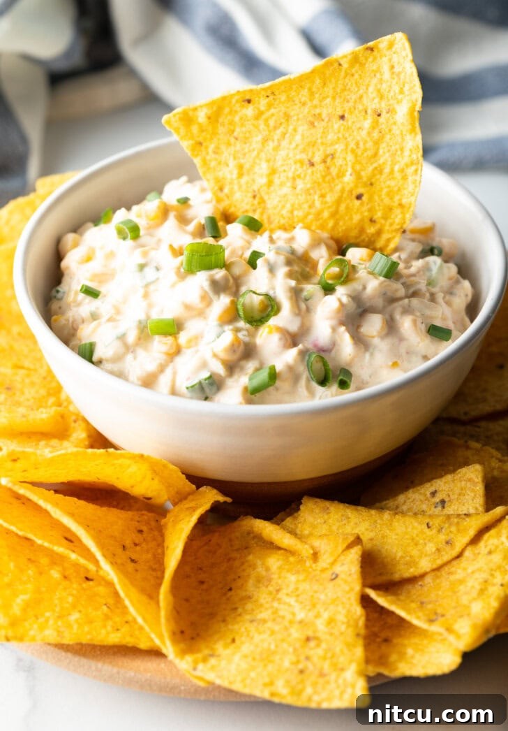 A tortilla chip laden with creamy Mexican corn dip being scooped from a pristine white bowl.