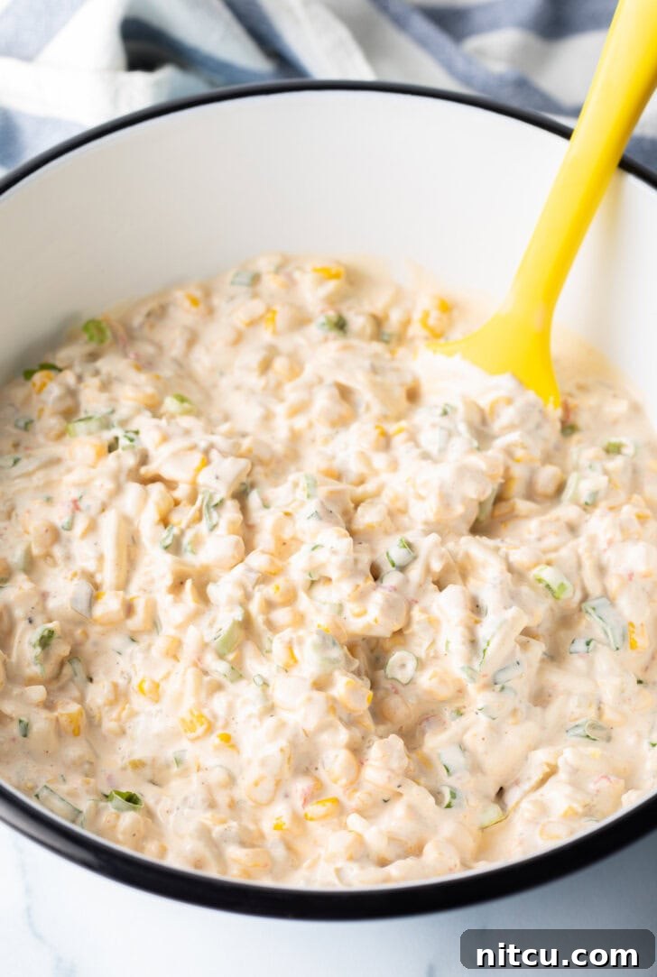 A white bowl of creamy corn dip being gently mixed with a yellow spatula, highlighting the smooth texture.