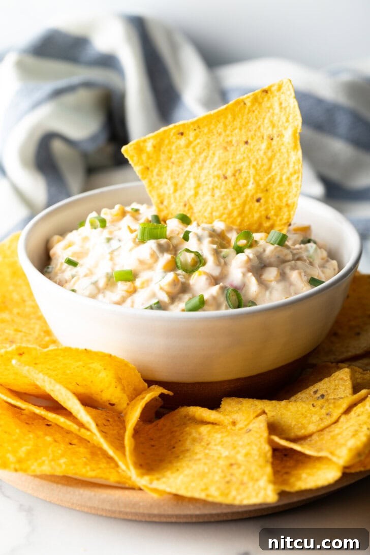 A tortilla chip loaded with creamy Mexican corn dip being scooped from a pristine white bowl, showcasing its rich texture.