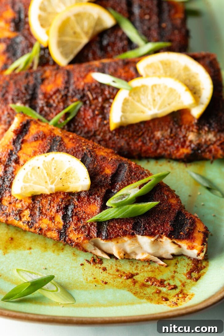 Close view of a fillet of grilled mahi mahi topped with a slice of lemon and green onions. A bite has been removed from the fish to show the white flaky inside of the meat.