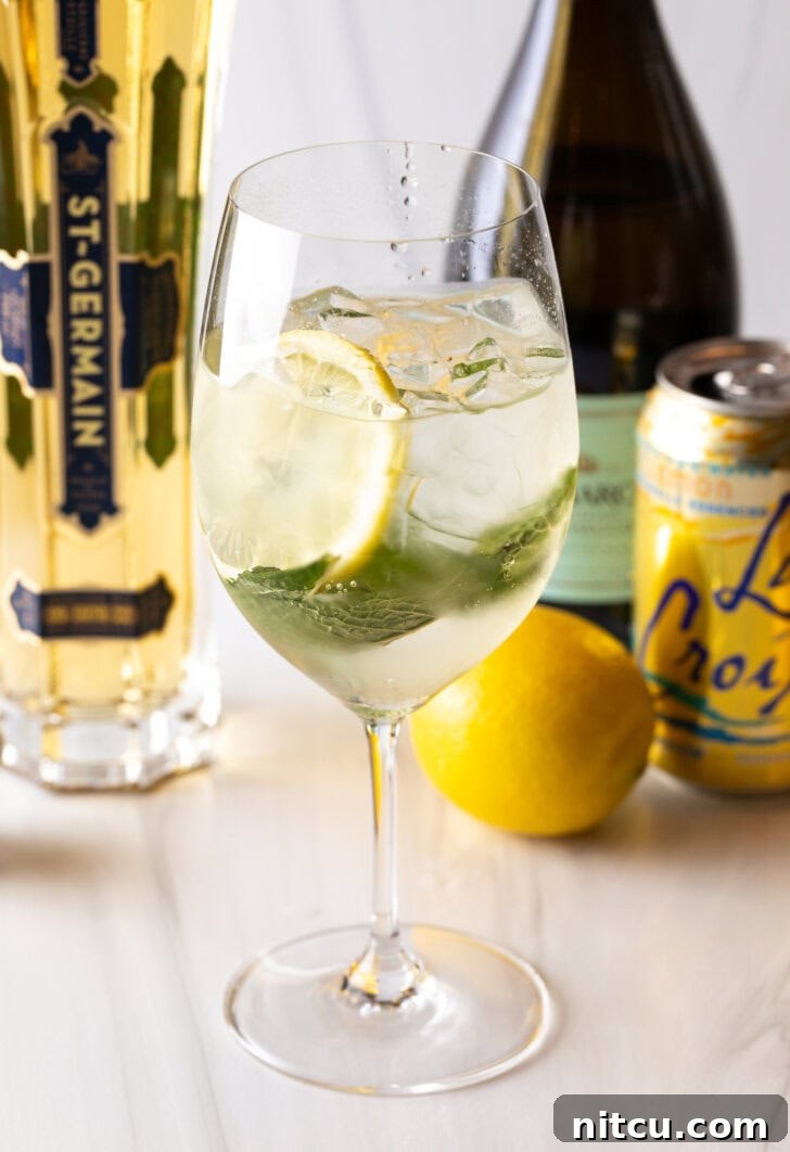 Wine glass with alcohol, mint leaves, and lemon slices. In the background of the hugo spritz are a whole lemon and bottles of prosecco, st. germain liquor, and a can of la croix soda.