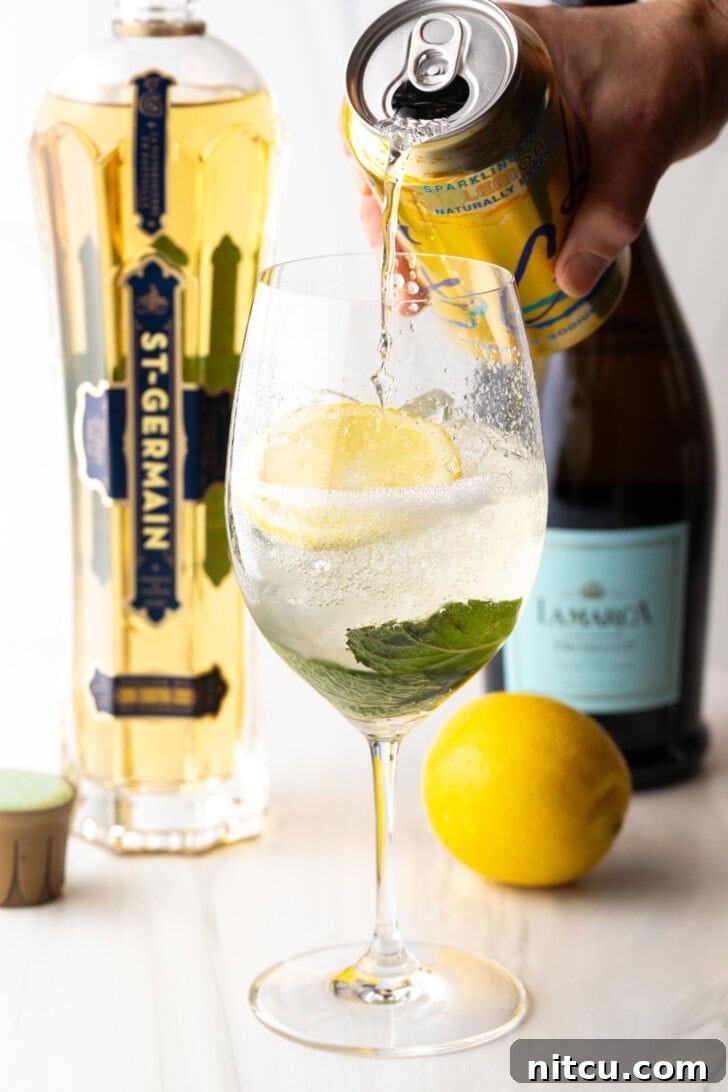 Wine glass with alcohol, mint leaves, and lemon slices. In the background are a whole lemon and bottles of champagne, st. germain liquor. A hand is adding soda from a can of la croix soda.