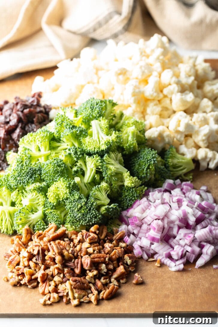 Cutting board showcasing neatly piled fresh broccoli florets, cauliflower florets, finely chopped red onions, crunchy pecans, and bright dried cranberries, ready for salad assembly.