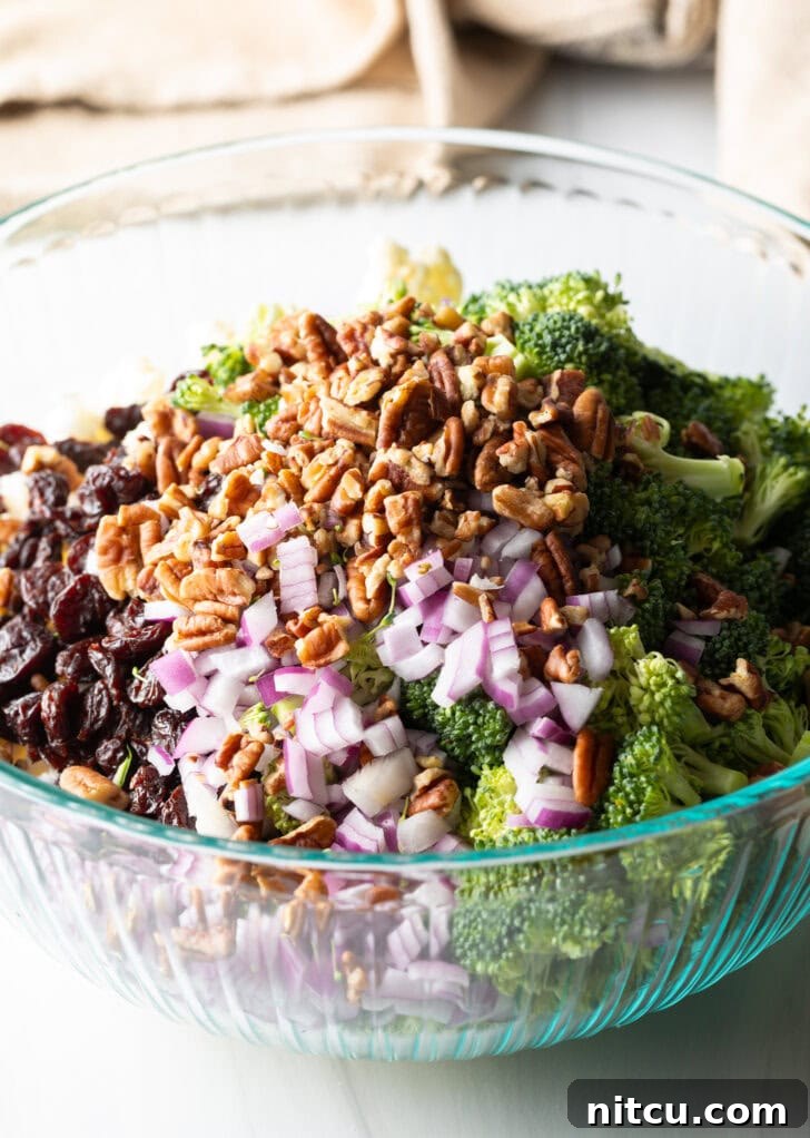 A large glass bowl filled with fresh broccoli florets, cauliflower florets, chopped red onions, pecans, and cranberries, awaiting the dressing.