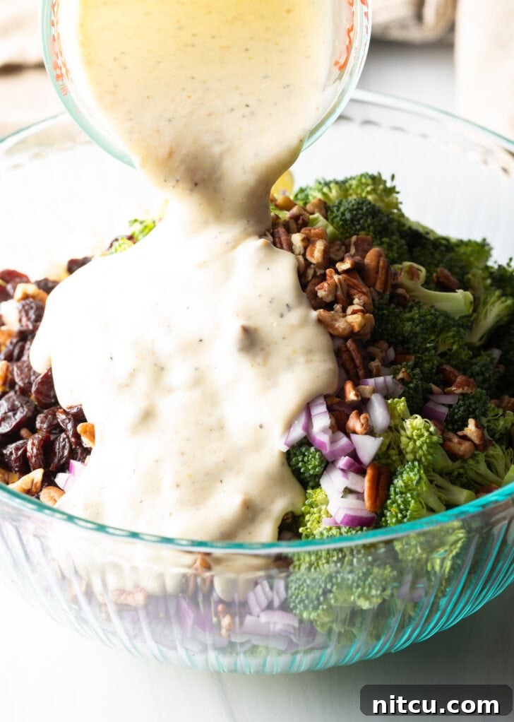 Creamy mayonnaise-based dressing being poured from a glass measuring cup onto the vibrant salad ingredients in a large glass bowl, illustrating the final step before tossing.
