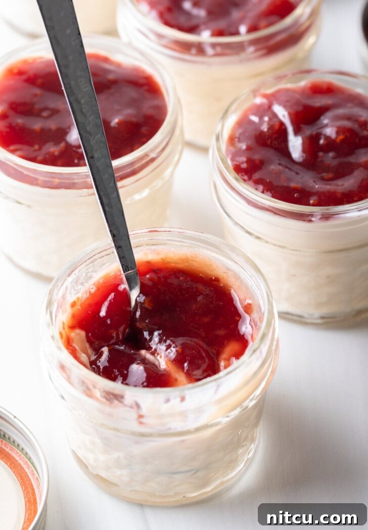 Top down view 4 small glass jars of protein pudding topped with raspberry jam. A metal spoon is in the jar closest to camera.