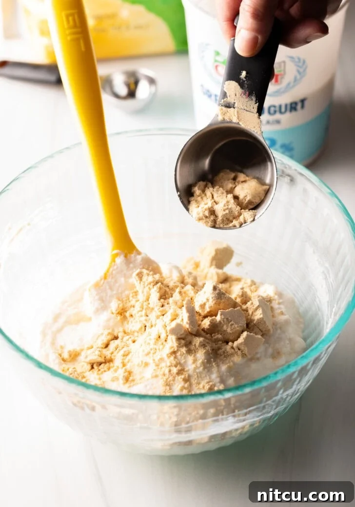 Adding protein powder from a metal measuring cup into a glass bowl with a yellow spatula.