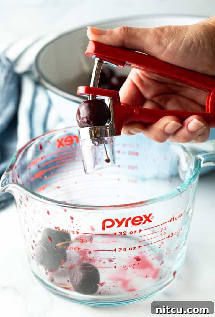 A hand skillfully uses a cherry pitter to remove the pit from a fresh cherry, illustrating a key preparation step for a homemade cherry pie.