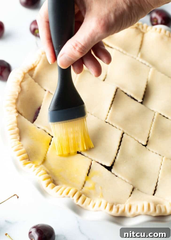 A hand gently brushes an egg wash over the intricate pattern of an unbaked pie's top crust, preparing it for a golden, glossy finish.