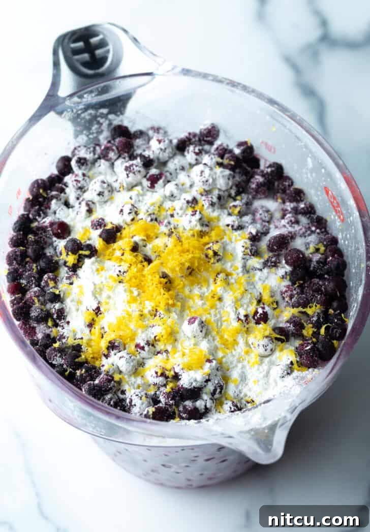 Close-up of a glass mixing bowl where fresh blueberries, lemon zest, sugar, and cornstarch are being gently combined with a spoon for the crisp filling.