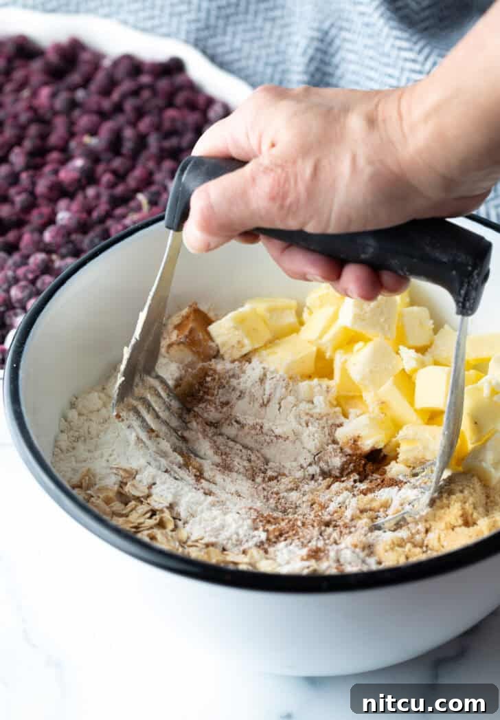 A close-up shot of a hand using a metal pastry cutter to cut cold butter into a mixture of flour, oats, and brown sugar in a bowl, creating a crumbly topping.
