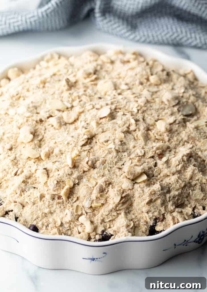 A baking dish filled with blueberries, covered generously with an uncooked, crumbly oat and almond topping, ready to be baked.