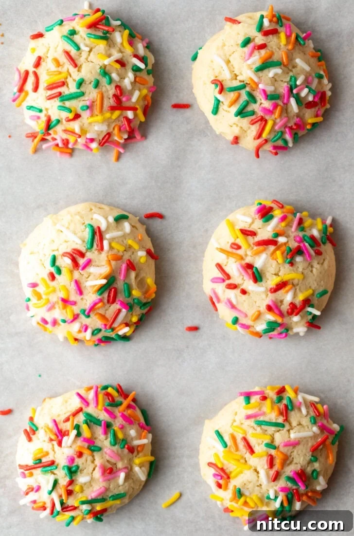Top down view 6 cream cheese cookies on a baking sheet, each topped with sprinkles.