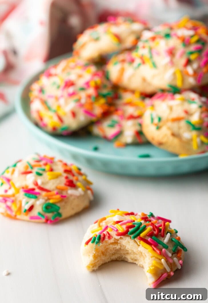 Cookie with sprinkles and a bit taken out is close to camera. Another cookie with no bite is off to the side. In the background is a plate piled up with cream cheese cookies.