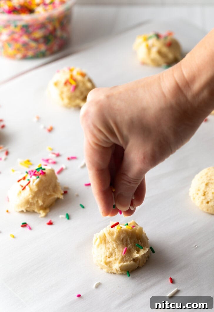 Hand adding colorful sprinkles to the tops of raw cookies balls spread out on a baking sheet.