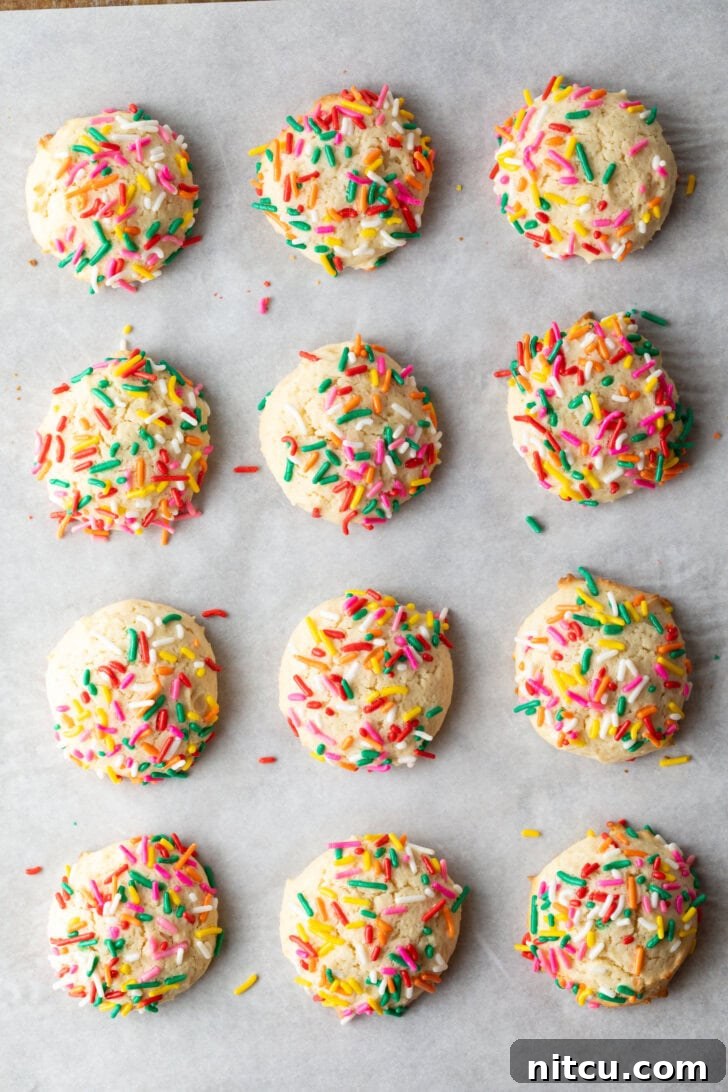 Top down view 12 cream cheese cookies on a baking sheet, each topped with sprinkles.